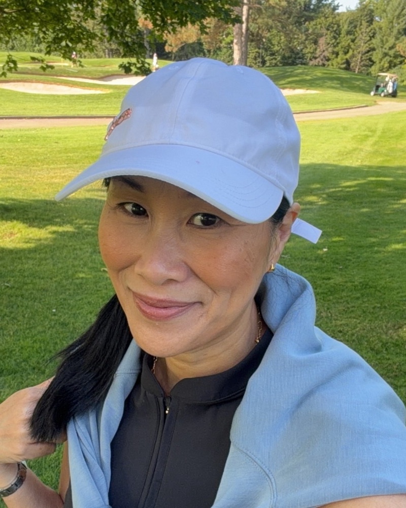 Suzan Chong smiling outdoors on a golf course, wearing a white cap